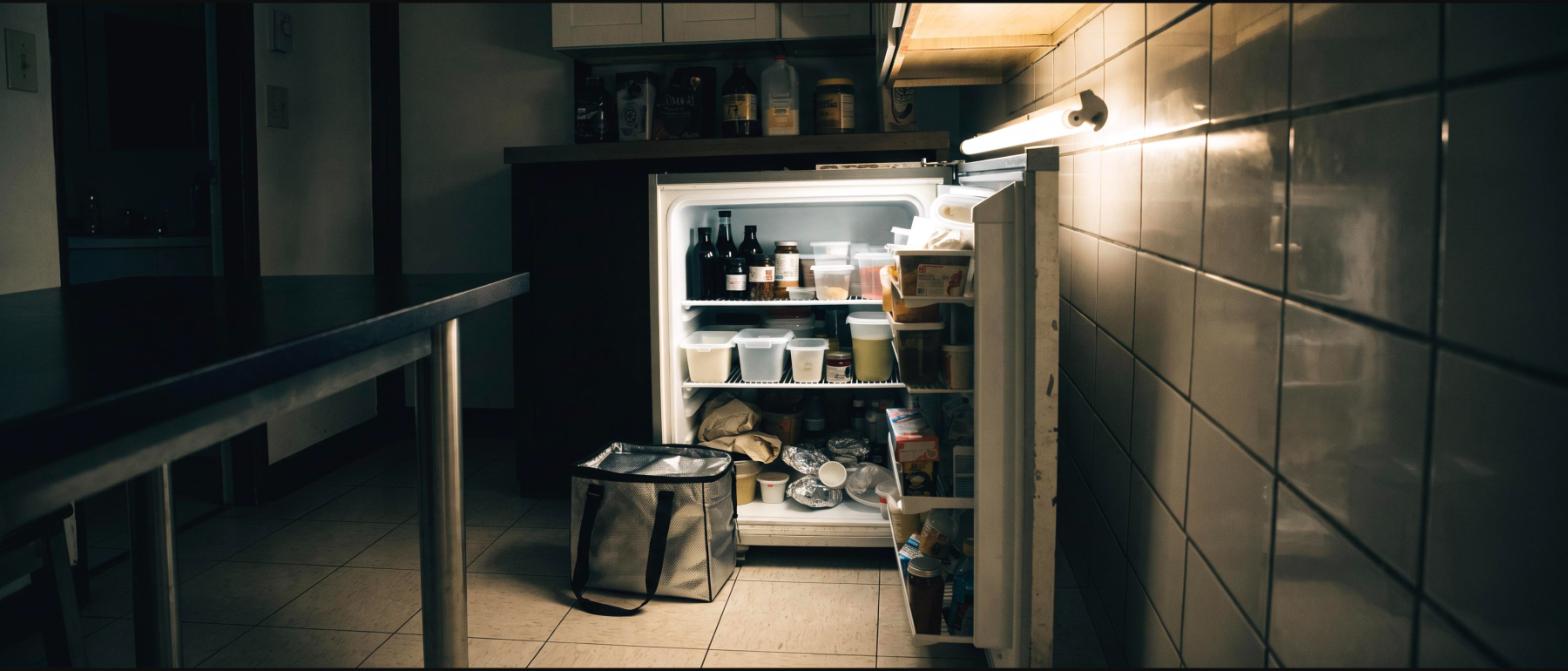 A small overcrowded office breakroom refrigerator standing open with no free shelf space and a lunch bag on the floor beside it